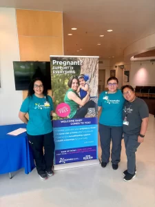 Three women stand by a banner that says "Pregnant? Support is Everything" with Emanate Health.